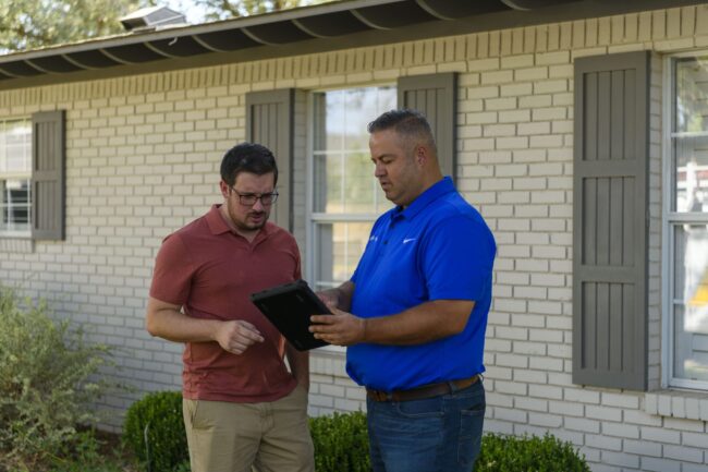 Two men discussing plumbing services outdoors, one holding a tablet, in front of a residential building with brick walls and shutters.
