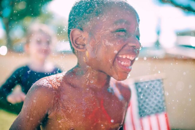 Happy child playing with water, enjoying summer fun outdoors, with a blurred background featuring an American flag.