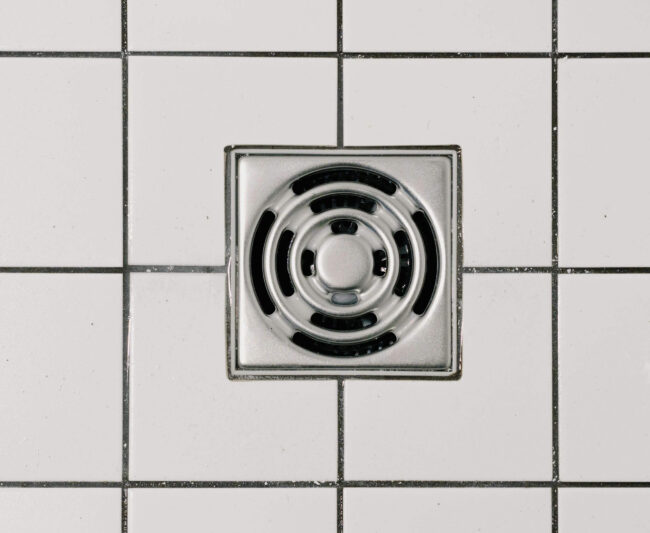 Silver floor drain cover on white tiled floor, relevant to plumbing services and hair-clogged drains.