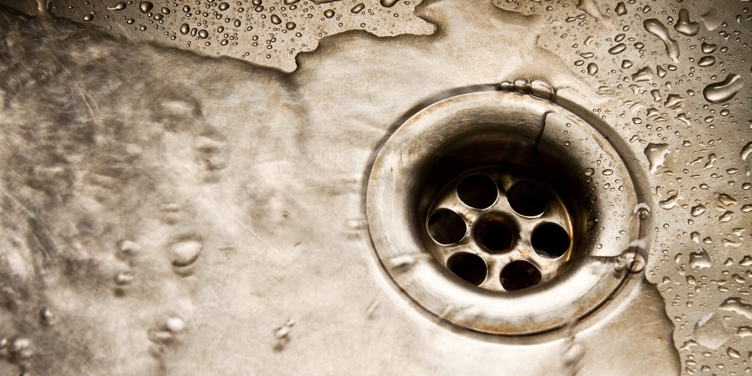 Close-up of a stainless steel sink drain surrounded by water droplets, illustrating potential plumbing issues like leaks or clogs relevant to plumbing services offered by I Fix Pipe.