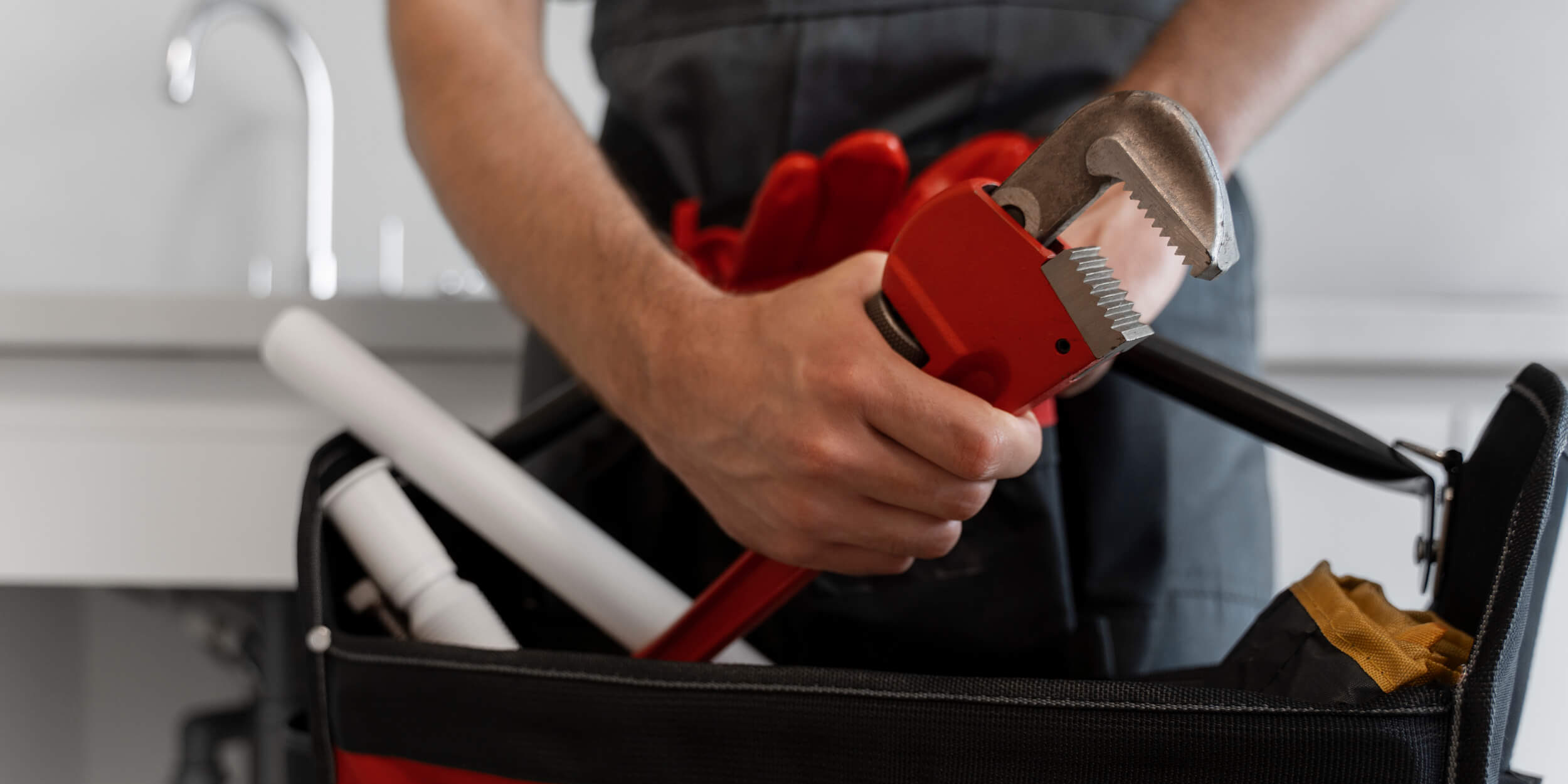 Man holding a red pipe wrench above a toolbox with plumbing supplies, symbolizing professional plumbing services.